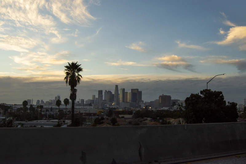 Downtown Los Angeles towers lit at dusk under a fading pink sky, seen from a freeway approach