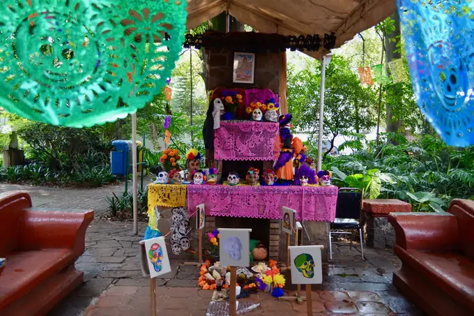 A tiered Día de Muertos altar draped in pink cut paper, set with sugar skulls, skeleton figures and orange marigolds under a garden canopy