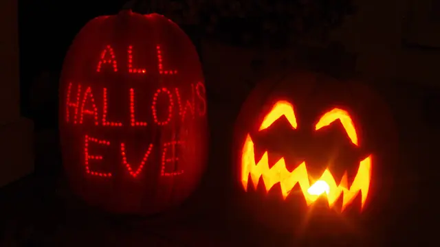 Two glowing jack-o’-lanterns at night, one carved with the words All Hallows Eve and one with a jagged grin