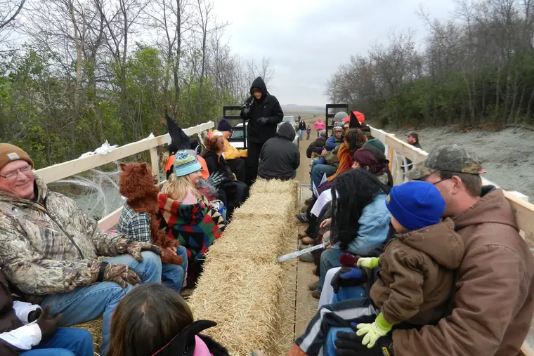 Adults and children, some in witch hats and costumes, ride on hay bales in a wagon along a wooded trail