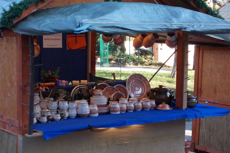 A wooden market stall with a blue counter cloth holds rows of handmade cream and brown pottery, with plates and pots hanging above