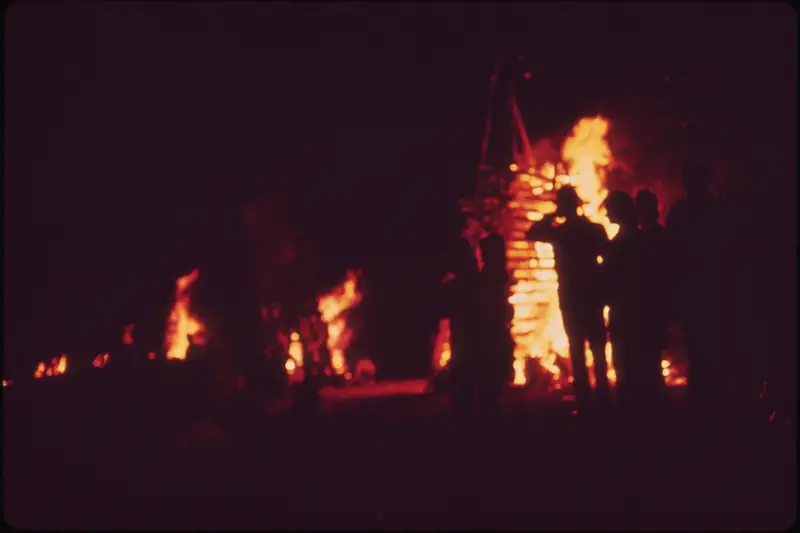 A tall wooden bonfire structure burning at night on a grassy Mississippi River levee