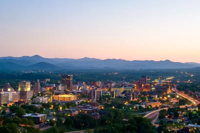 Downtown Asheville buildings and streets lit at dusk, with layered blue mountains under a pink sky