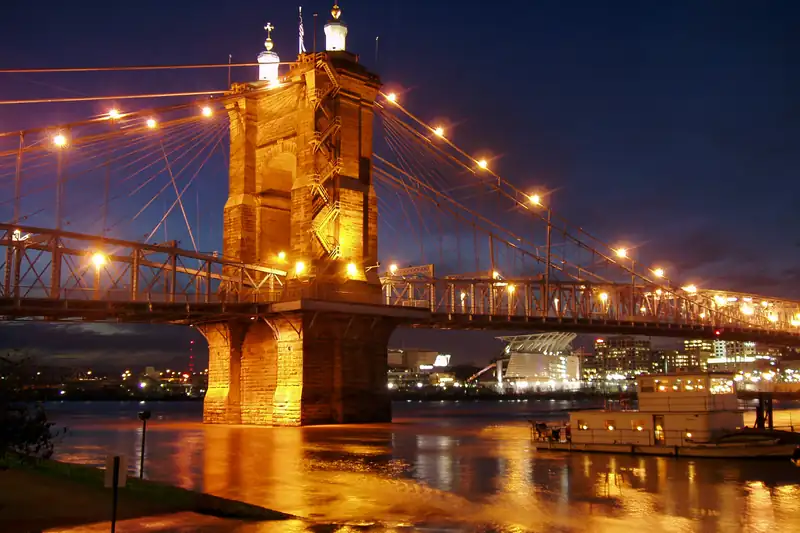 The stone tower and lit cables of a suspension bridge over a wide river at dusk, with city lights on the far bank
