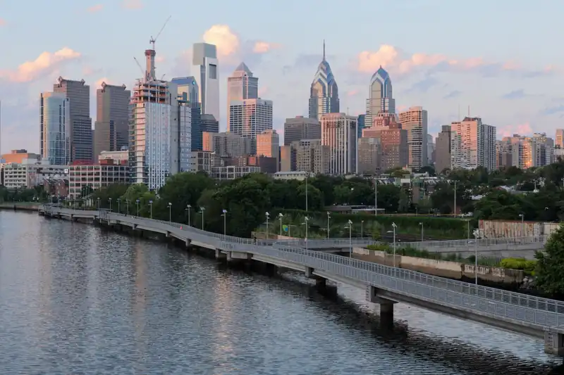 Philadelphia downtown towers above green trees along the Schuylkill River, with a riverside boardwalk trail curving along the water at dusk