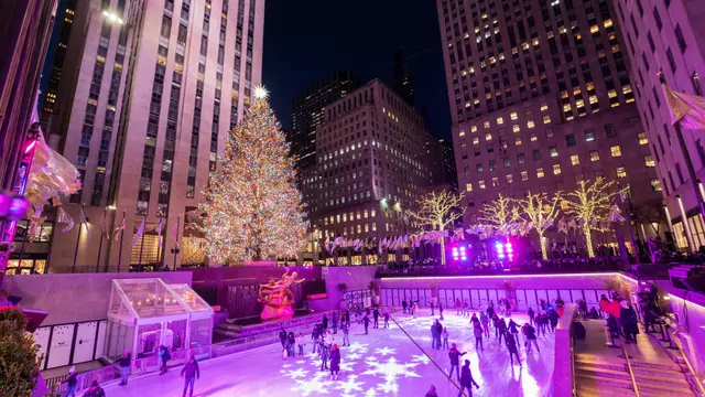 A tall Christmas tree covered in colored lights above a crowded ice rink between skyscrapers at night