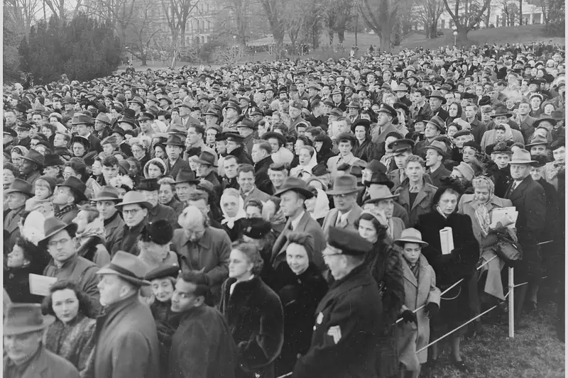 Black-and-white photo of a large crowd in hats and winter coats gathered for a Christmas tree lighting near the White House