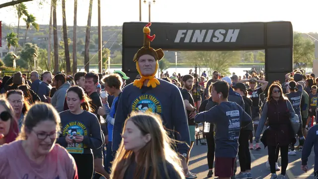 Runners and walkers in race shirts near a finish arch at a turkey trot, one man wearing a turkey hat