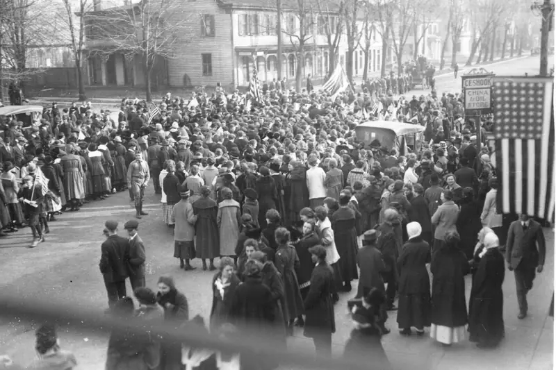 Black-and-white photo of a large crowd filling a small-town street on Armistice Day 1918, with an American flag hanging at right