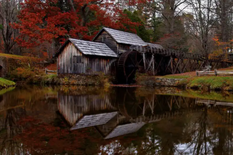 A wooden gristmill with a water wheel reflected in a still pond, under red and orange autumn trees